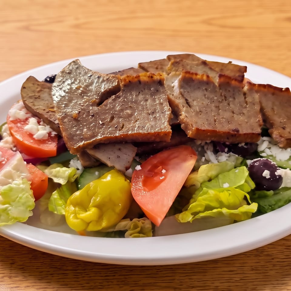 A gyro salad with sliced meat, lettuce, tomatoes, feta cheese, olives, and peppers on a white plate.
